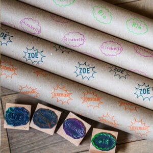 Four Custom Comic Kids Name Rubber Stamps next to four rolls of kraft paper stamped with four different custom kids name stamps